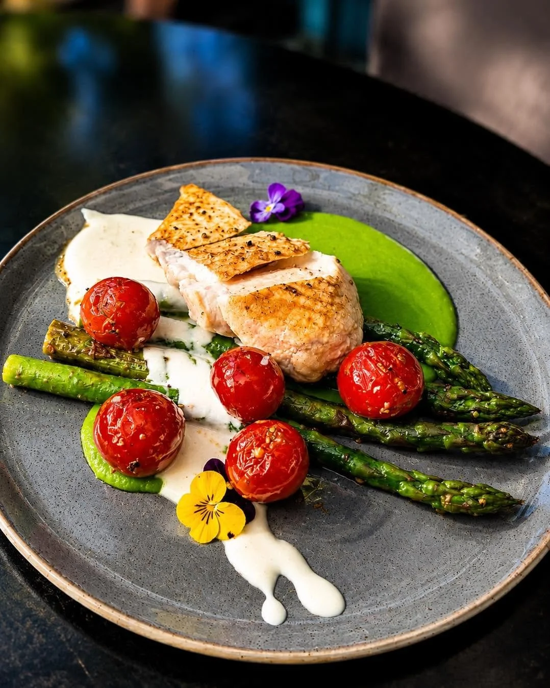 A plate of cooked salmon, roasted cherry tomatoes, grilled asparagus, green puree, and garnished with edible flowers and white sauce.