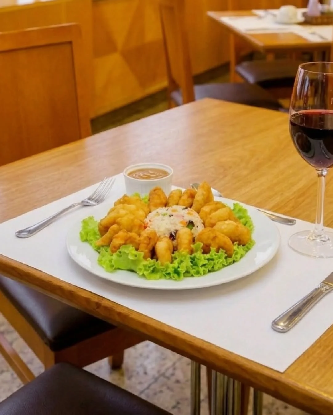 Plate of assorted fried seafood, including shrimp and fish, garnished with lettuce, accompanied by a small bowl of dipping sauce, a glass of red wine, and a roll of sushi, set on a white placemat on a wooden table in a restaurant.