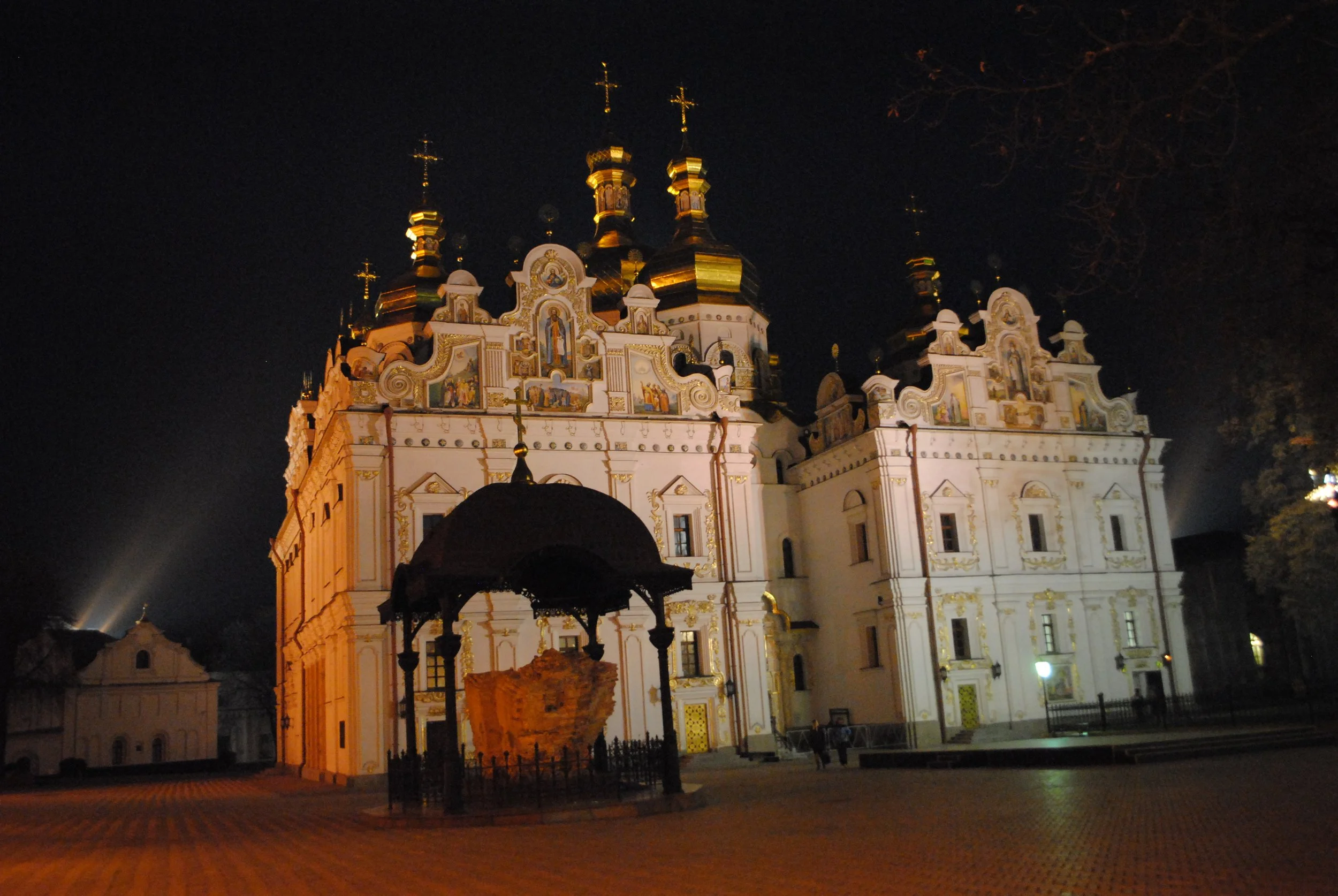 Night view of a brightly illuminated church with golden domes and crosses, visible from a courtyard with brick pavement and lanterns.