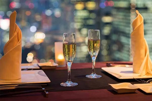 A romantic dining setup with two glasses of champagne, yellow folded napkins, plates, and candles at an elegant restaurant with city lights in the background.