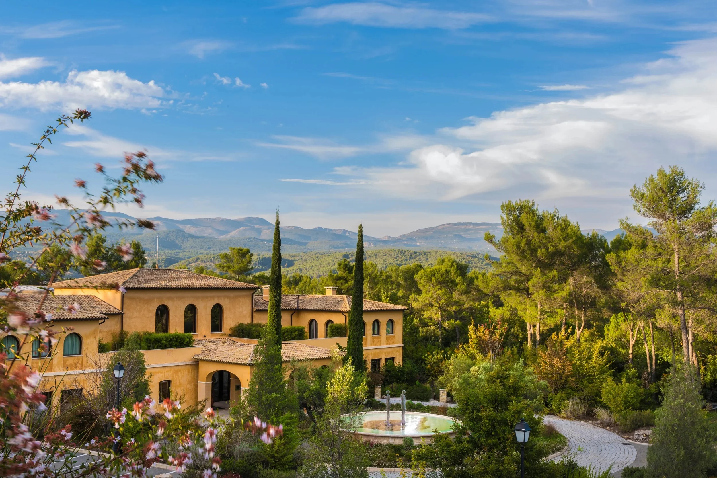 A scenic view of a Mediterranean-style estate surrounded by lush greenery, tall cypress trees, and mountains in the distance under a partly cloudy sky.