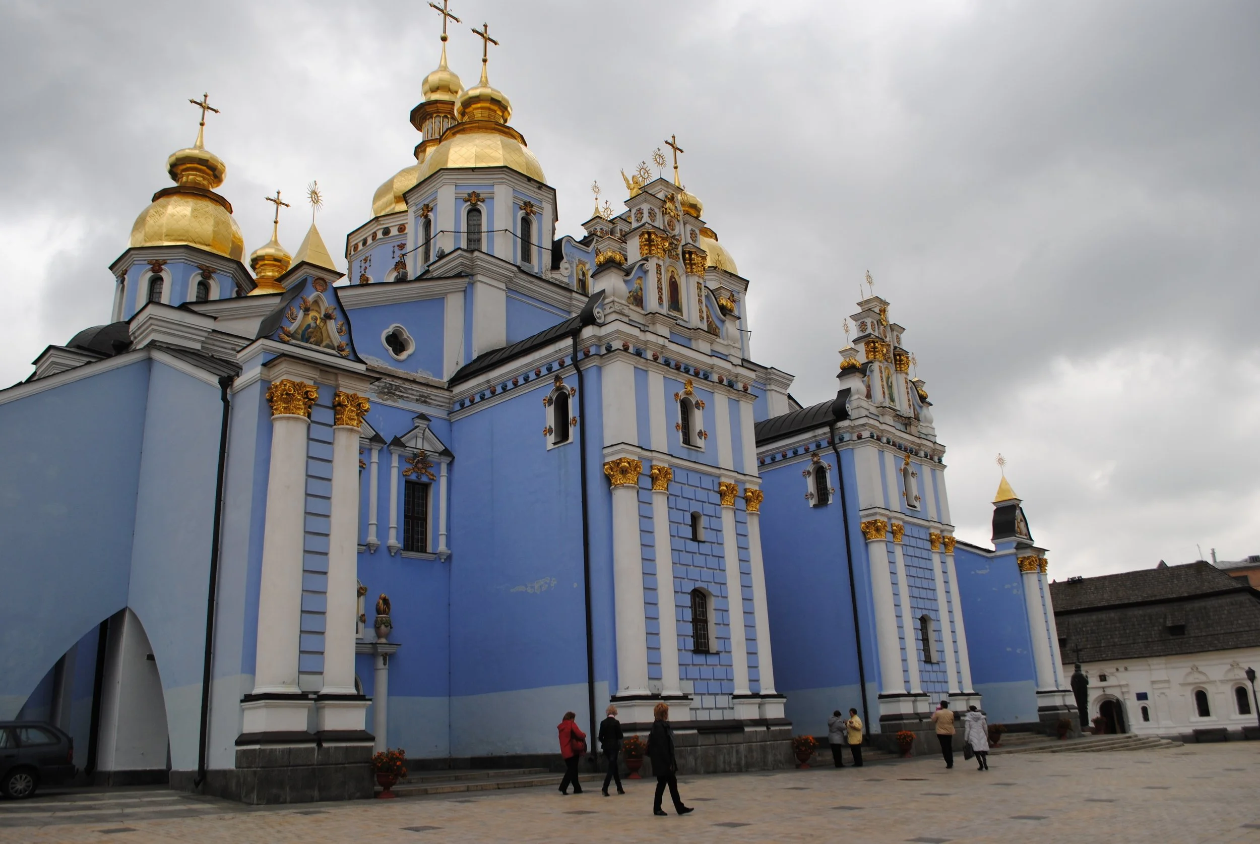A large, ornate church with blue and white walls and golden domes topped with crosses. Several people are walking near the church, and the sky is overcast with dark clouds.