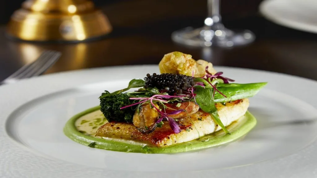 A gourmet dish with a piece of white fish, green vegetables, purple microgreens, caviar, and a green sauce on a white plate.