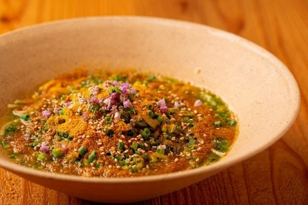 Soup in a beige bowl topped with chopped herbs, sesame seeds, and a pink flower-shaped garnish, on a wooden table.