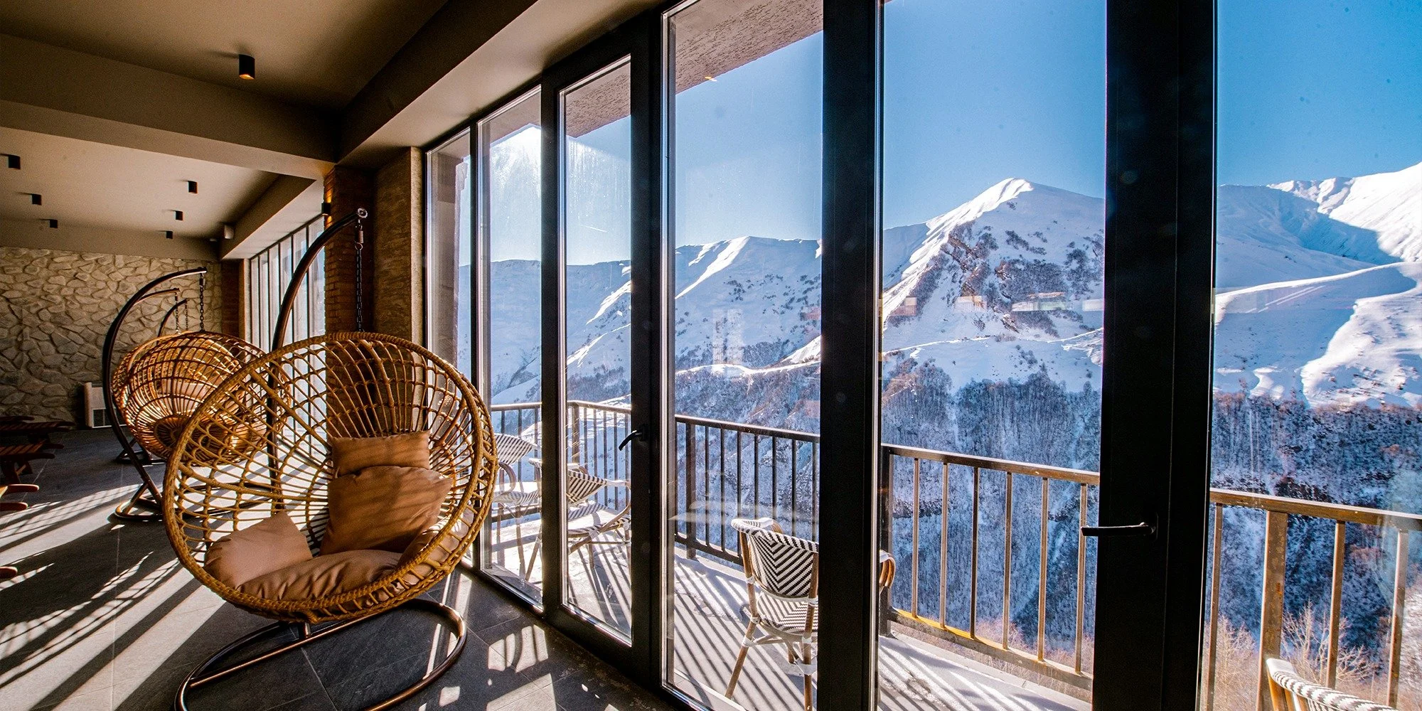 Interior of a room with large glass windows overlooking snow-covered mountains and a ski resort, featuring hanging wicker chairs with cushions, a stone wall, and a balcony.