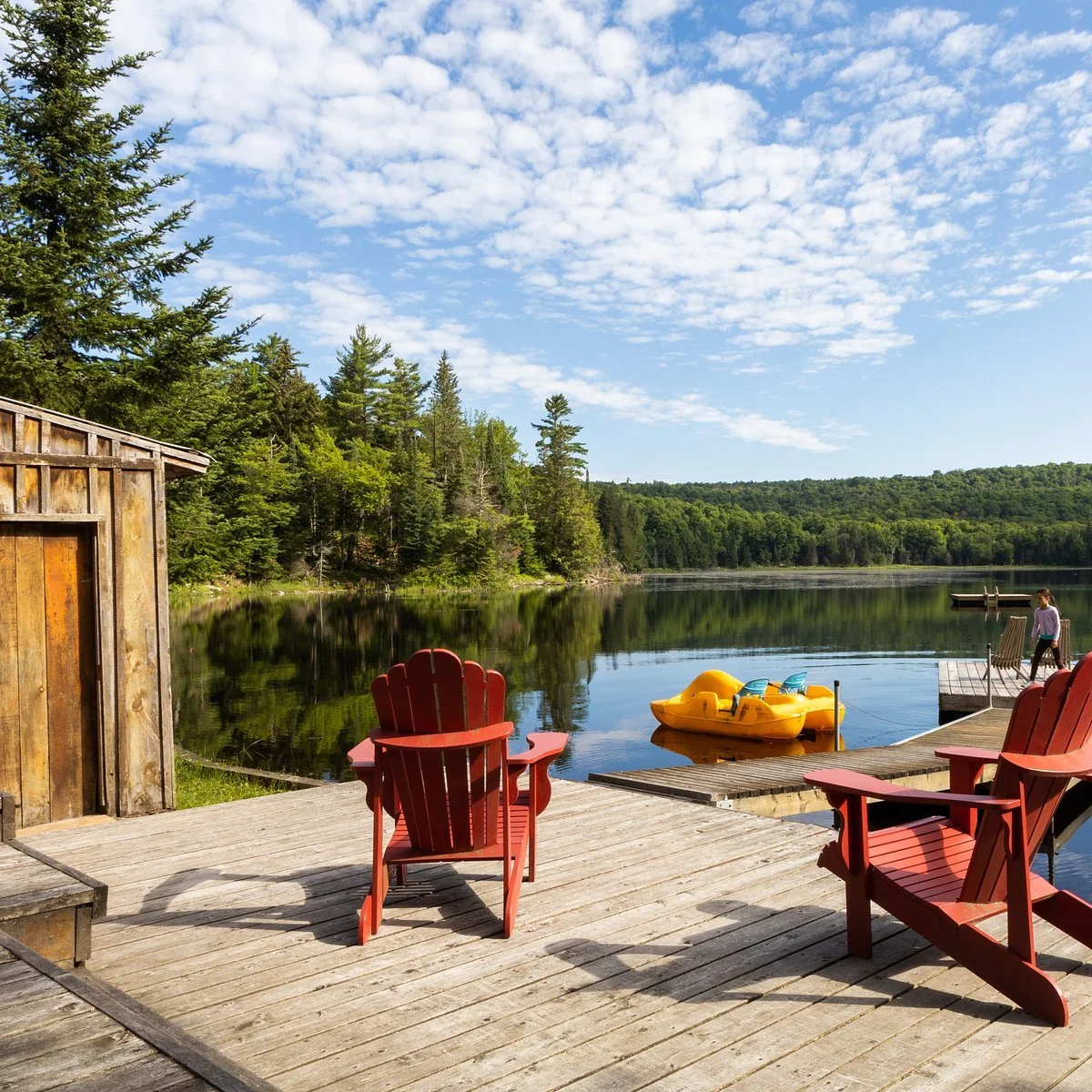 A lakeside scene with red Adirondack chairs on a wooden dock, a yellow paddleboat on the water, surrounded by green trees and a blue sky with scattered clouds.