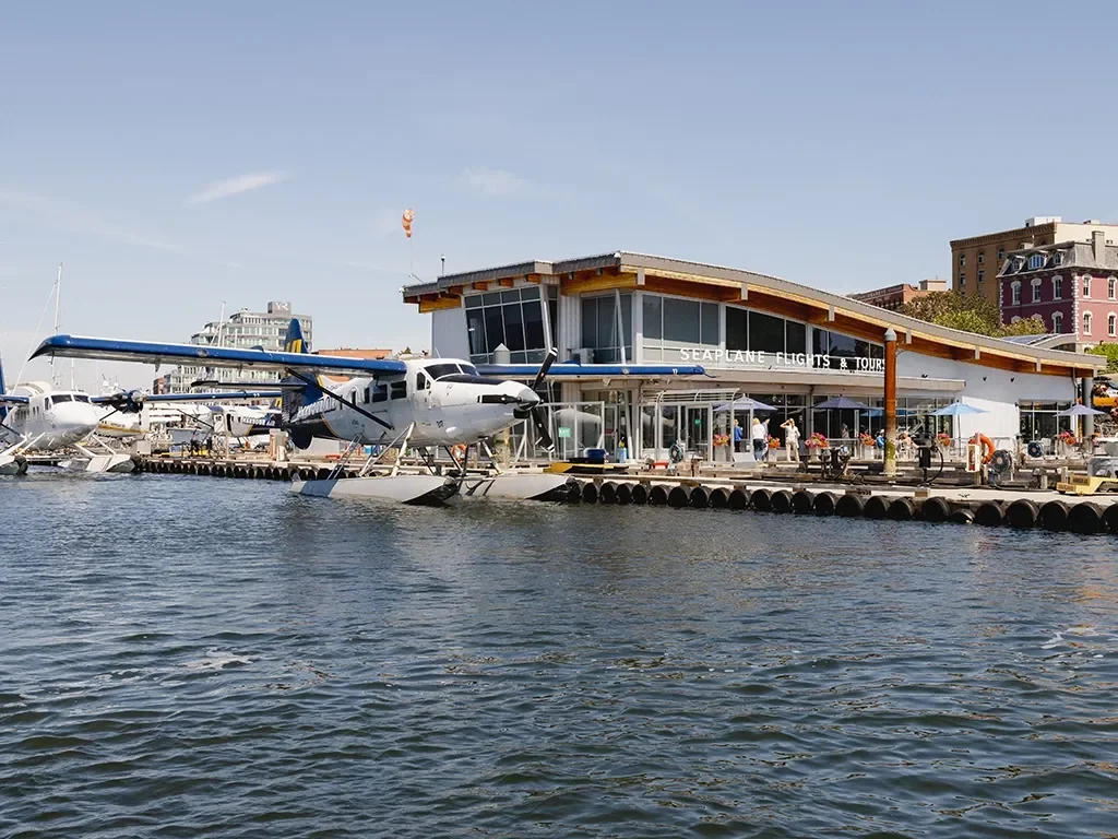 Seaplane docked at a waterfront with parked boats and a modern building in the background.