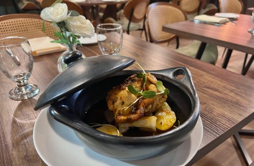 Dinner table with a plate of roasted chicken and potatoes in a black dish, surrounded by glasses, plates, utensils, and a small white flower bouquet in a restaurant setting.