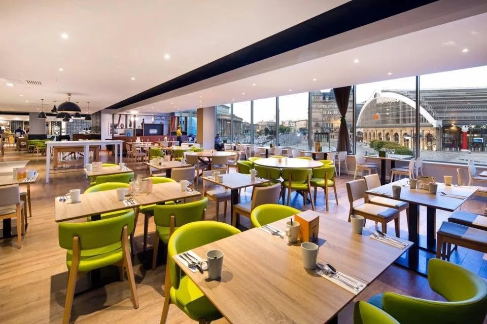 Bright, modern restaurant with wooden tables and green and beige chairs, large windows with a city view, and groups of cups and cutlery set on tables.