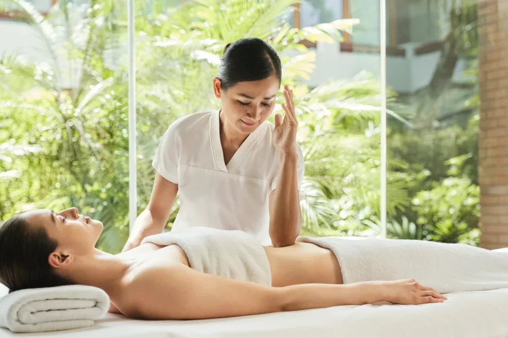 Spa therapist giving a facial massage to a woman lying on a massage table in a bright, natural-lit spa setting with green plants outside the window.