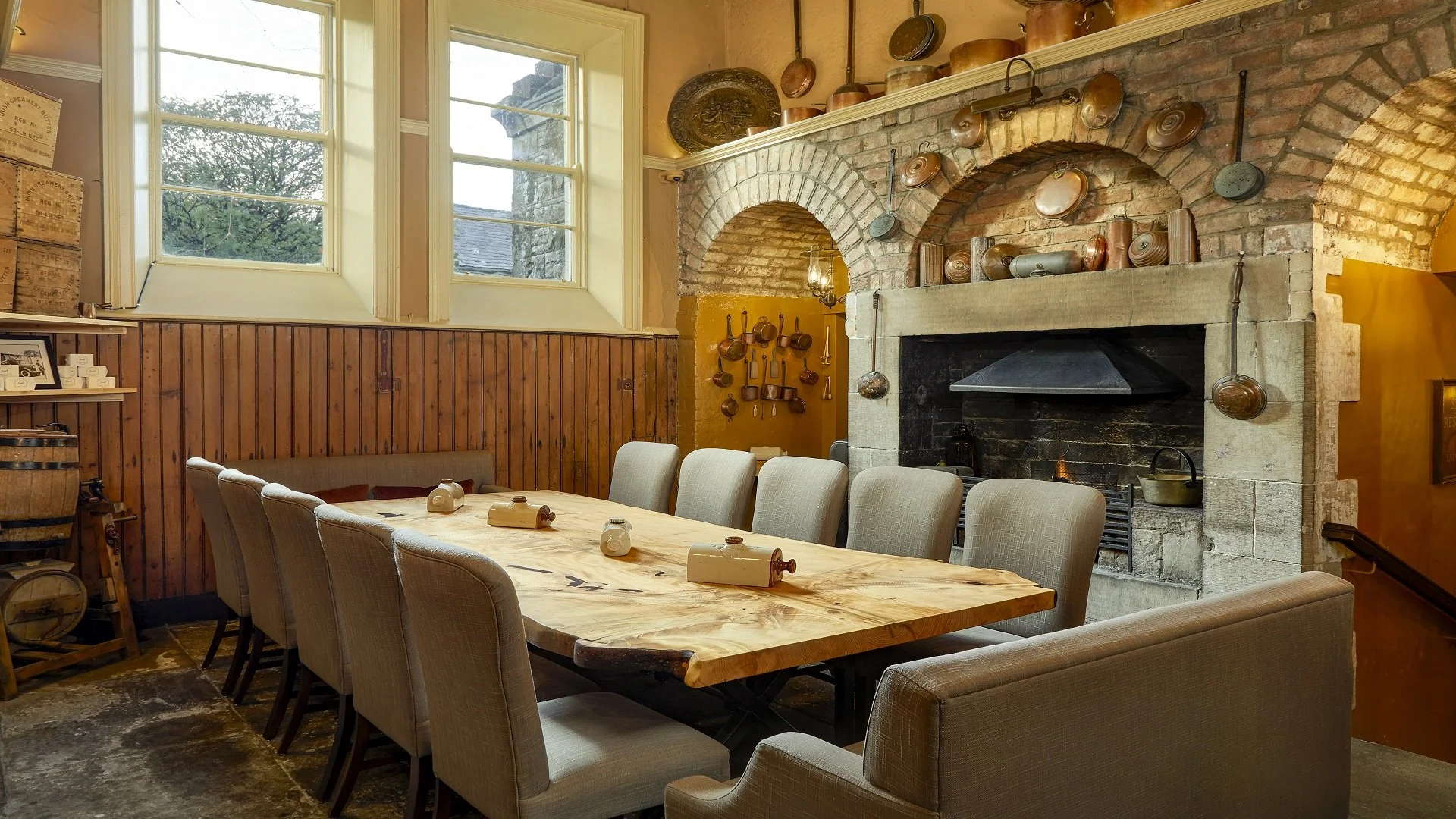 A cozy dining room with a large wooden table surrounded by beige upholstered chairs. The room features a brick fireplace with a rustic mantel adorned with copper pots and pans. Two large windows let in natural light and reveal trees outside. The wall