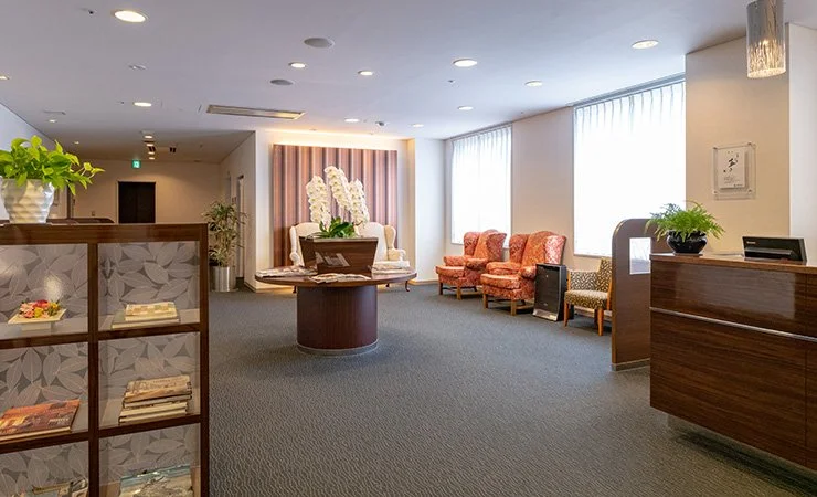 Lobby area with seating, a round table with a flower arrangement, and a reception desk. Bright and welcoming with patterned armchairs, large window letting in natural light, and decorative accents.