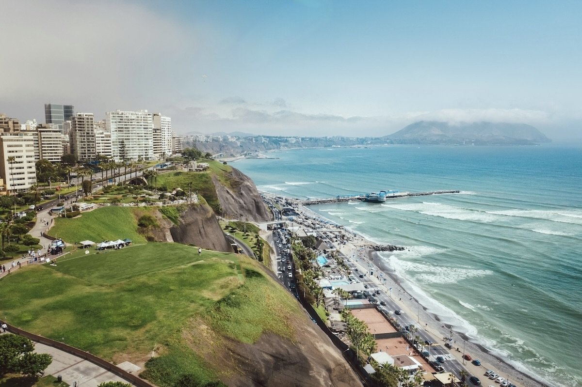 Coastal cityscape with high-rise buildings on cliffs, a green park area, a busy road below, and a beach with a pier extending into the ocean under a partly cloudy sky.