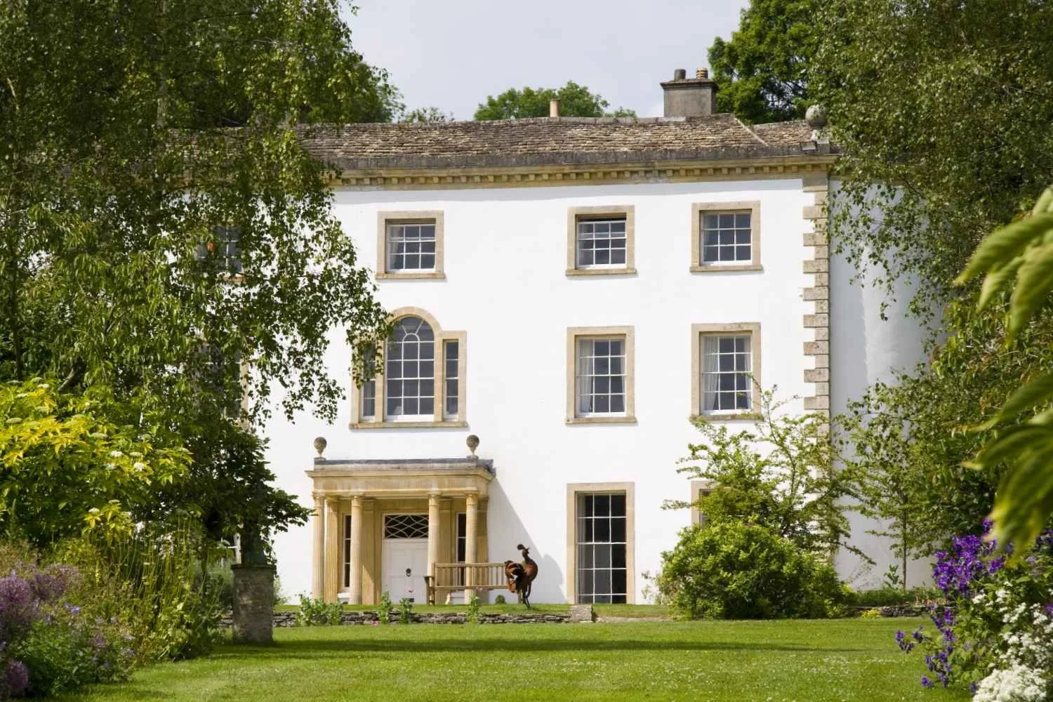 A white, three-story house with large windows, a small porch with columns, surrounded by green trees and a well-maintained lawn, with a decorative sculpture near the entrance.