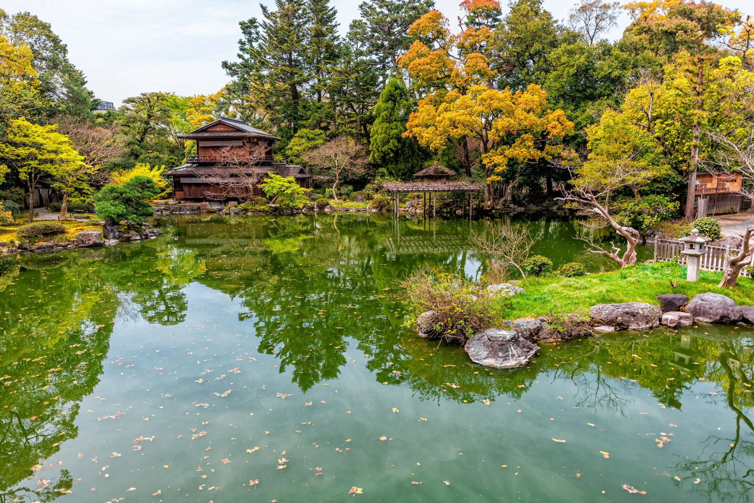 A traditional Japanese garden with a pond, surrounded by trees with green and yellow leaves, and wooden structures including a pagoda and a small bridge.