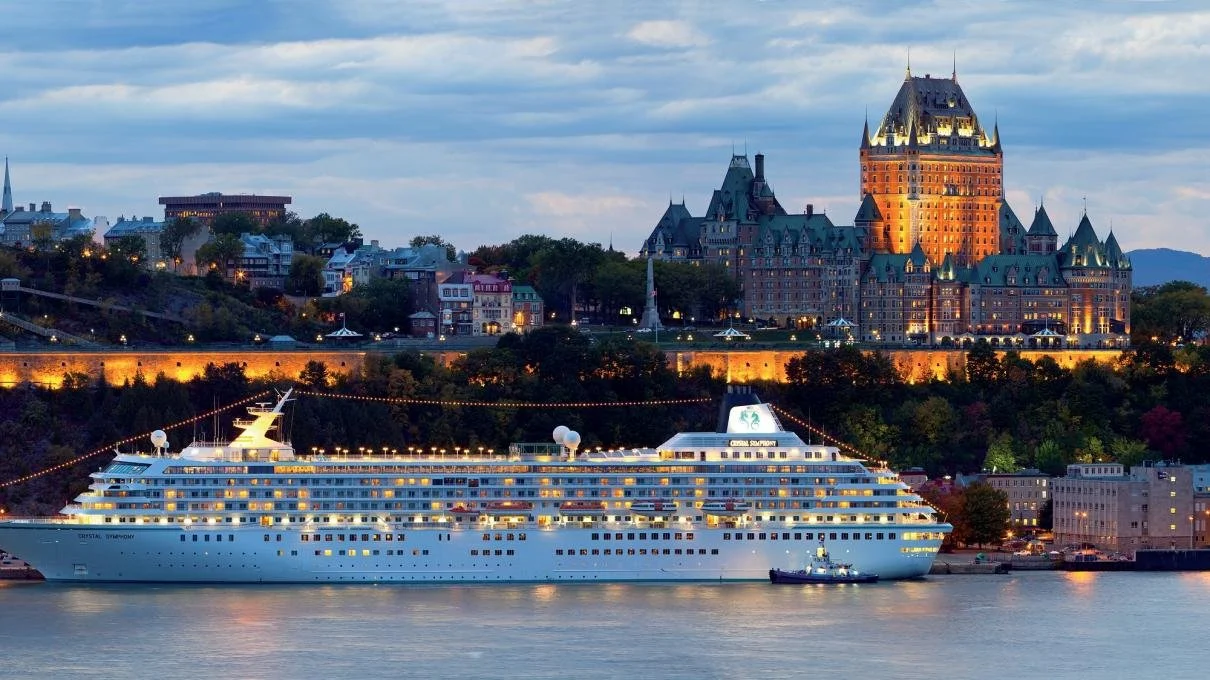 A large cruise ship docked on a river with a historic castle-like hotel in the background, illuminated at dusk.