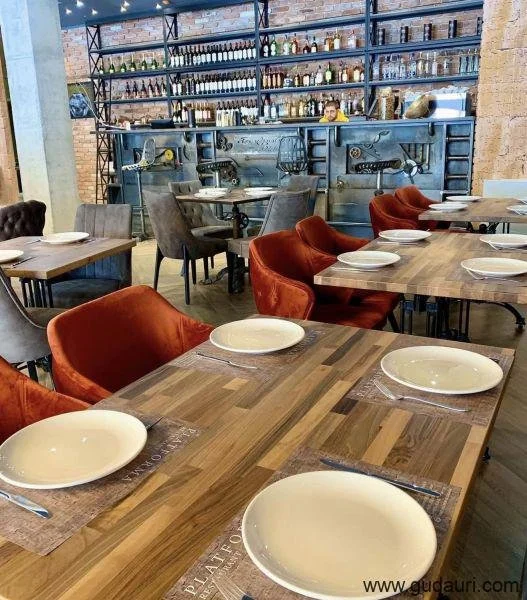 Empty dining tables with white plates and silverware in a restaurant, with a bar in the background and colorful upholstered chairs surrounding the tables.