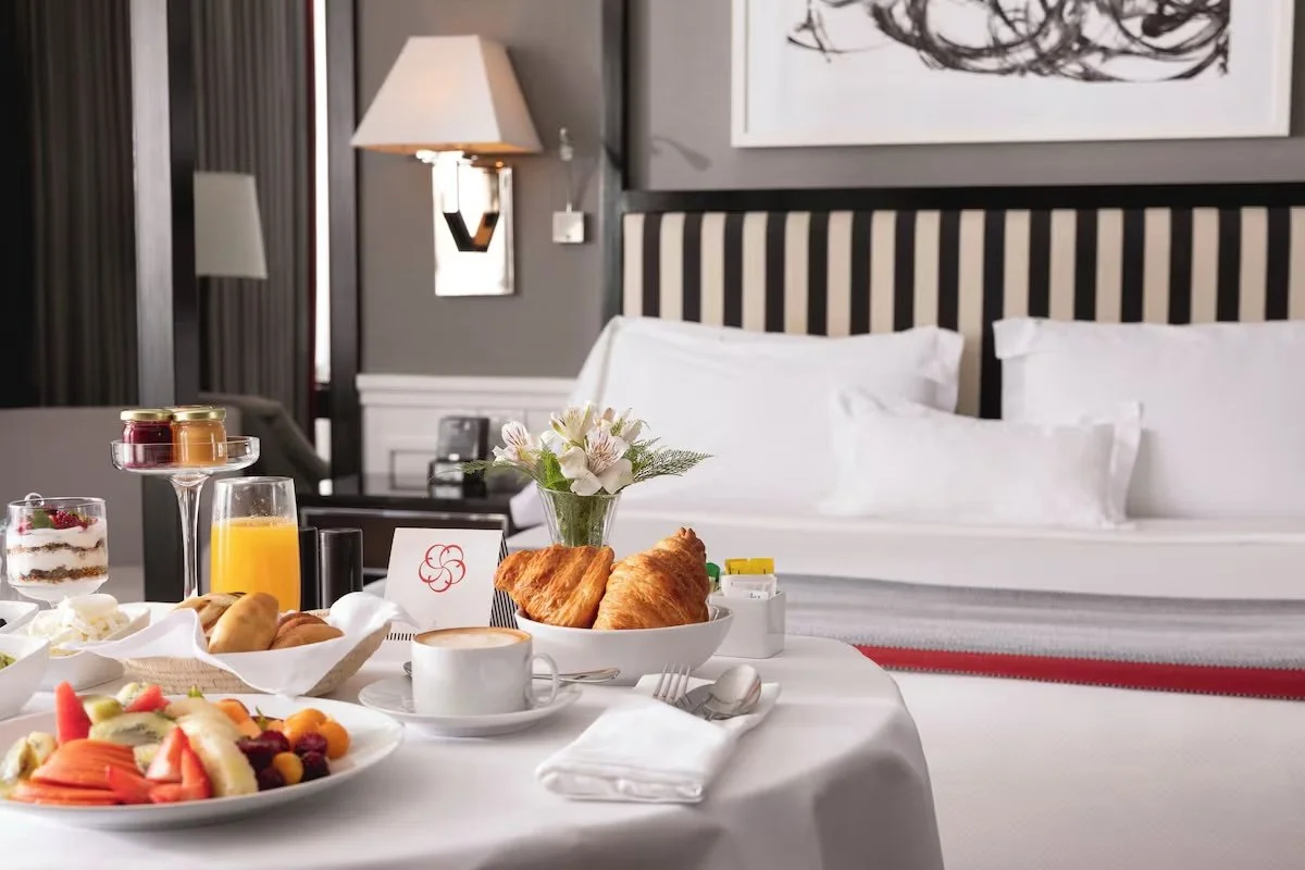 Breakfast spread on a hotel room table with croissants, assorted fruits, yogurt, orange juice, coffee, and a small flower arrangement against a background of a bed with white pillows and a striped headboard, with wall art and lamps in the room.