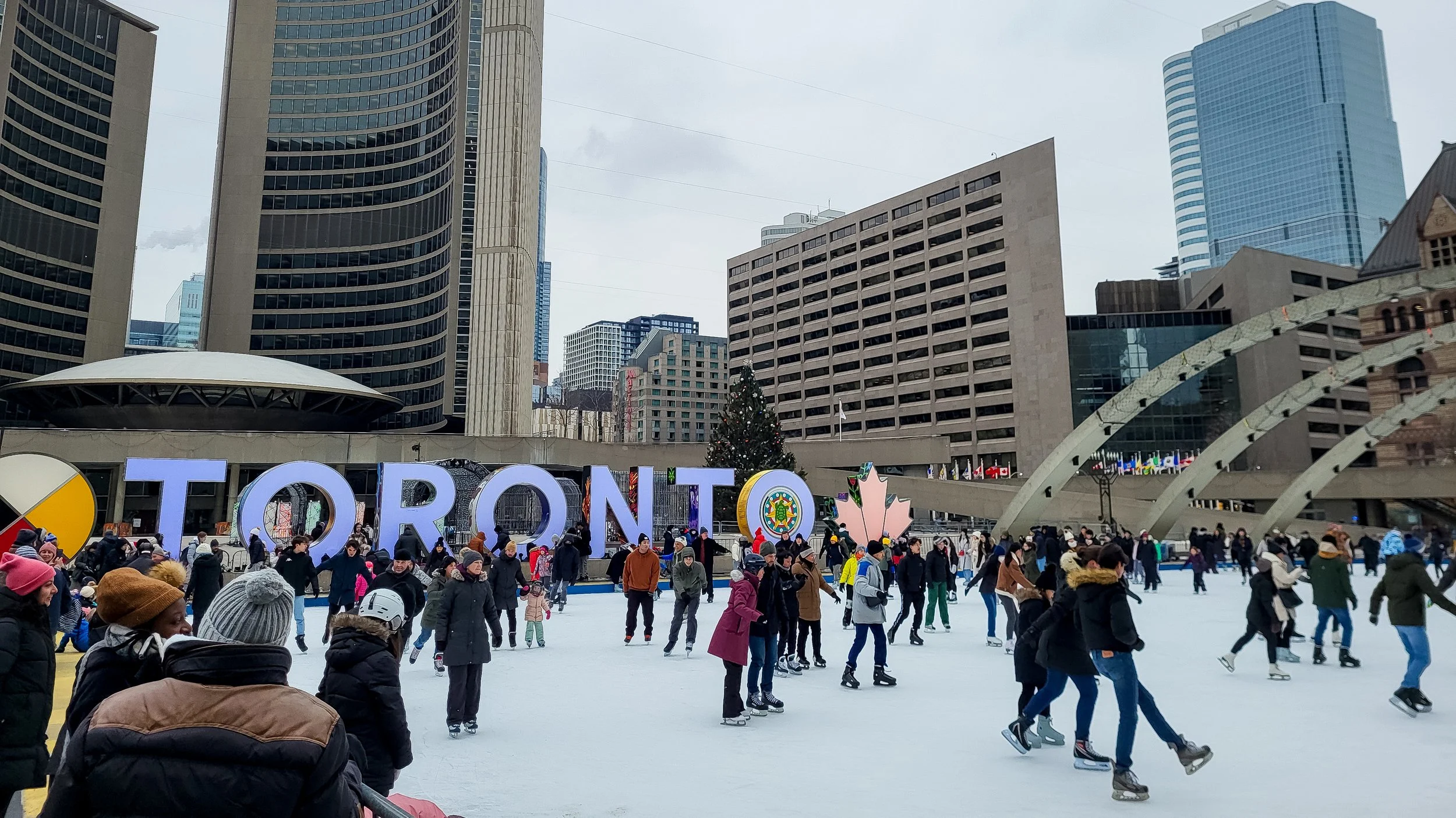 People ice skating on an outdoor rink in Toronto with large colorful 'TORONTO' sign and city skyscrapers in the background.