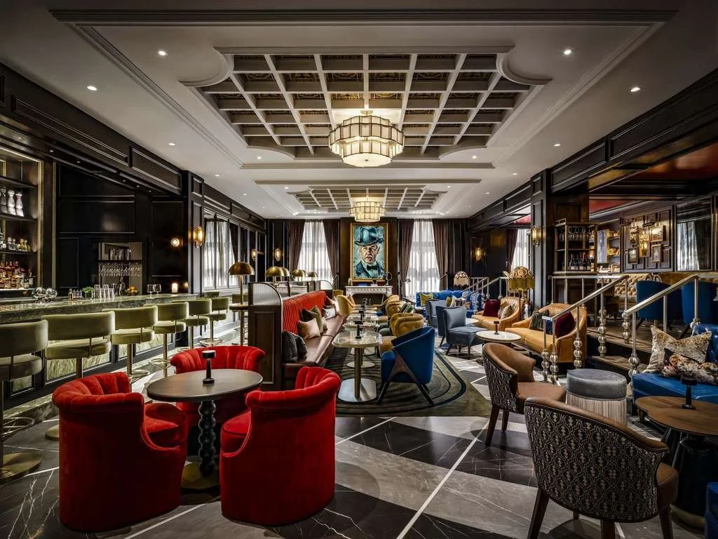 A luxurious lounge with colorful seating, dark wood paneling, large windows with curtains, and a central ceiling light fixture. There is a bar counter on the left and a large portrait painting on the back wall.