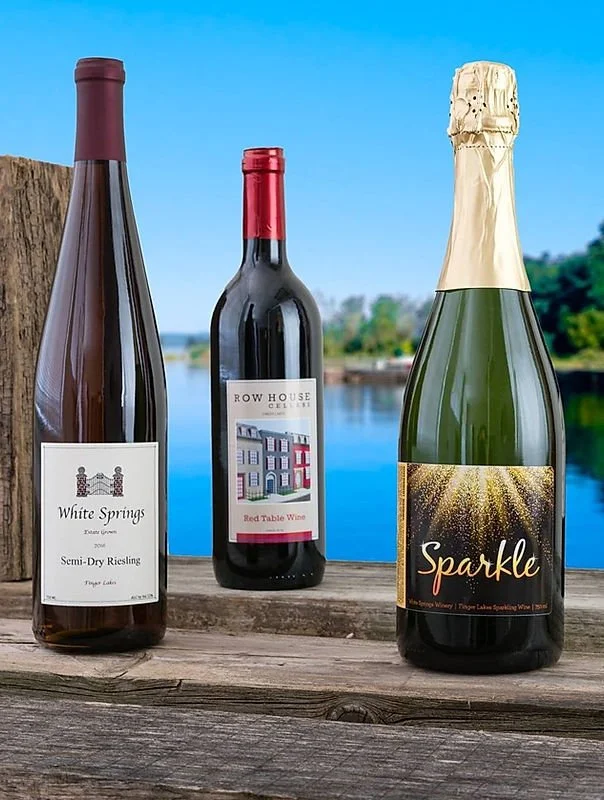 Three bottles of wine placed on a wooden surface outdoors near a body of water with trees in the background. The first bottle on the left is a semi-dry Riesling from White Springs, the middle is a red wine called Row House Cellars, and the right bottle is sparkling wine labeled Sparkle.