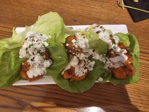 Three stuffed lettuce wraps with fried chicken, blue cheese crumbles, and a drizzle of dressing on a white rectangular plate.