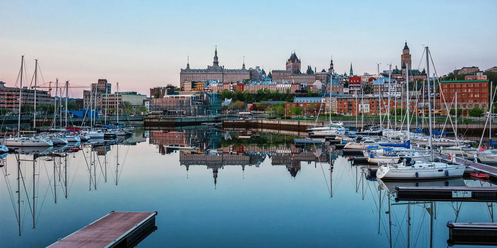 A marina with sailboats docked in calm water and a city skyline behind, including historic and modern buildings on a hill, with reflections visible in the water.
