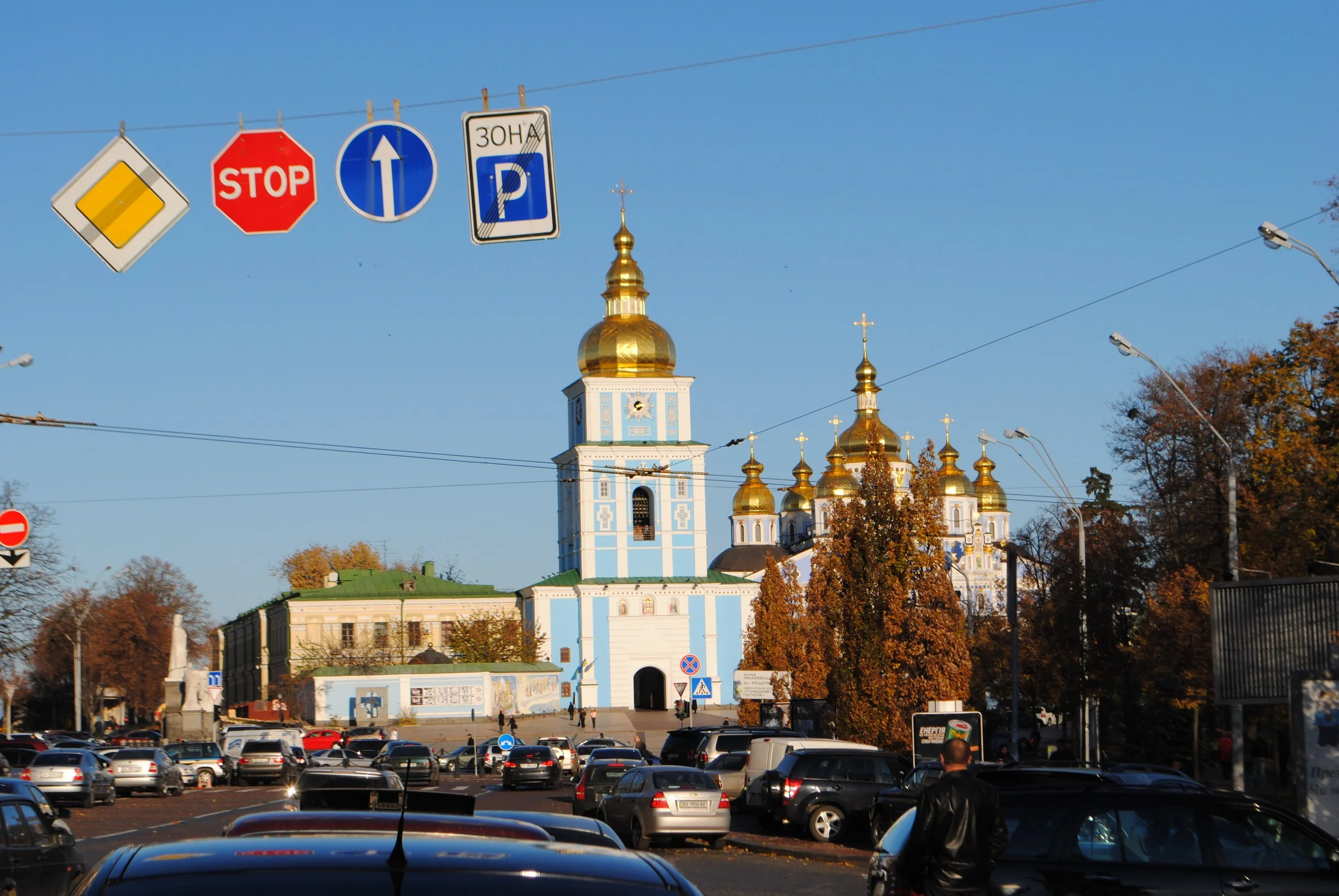 City square with parked cars in the foreground, a large blue and white church with golden domes in the background, and a clear blue sky.