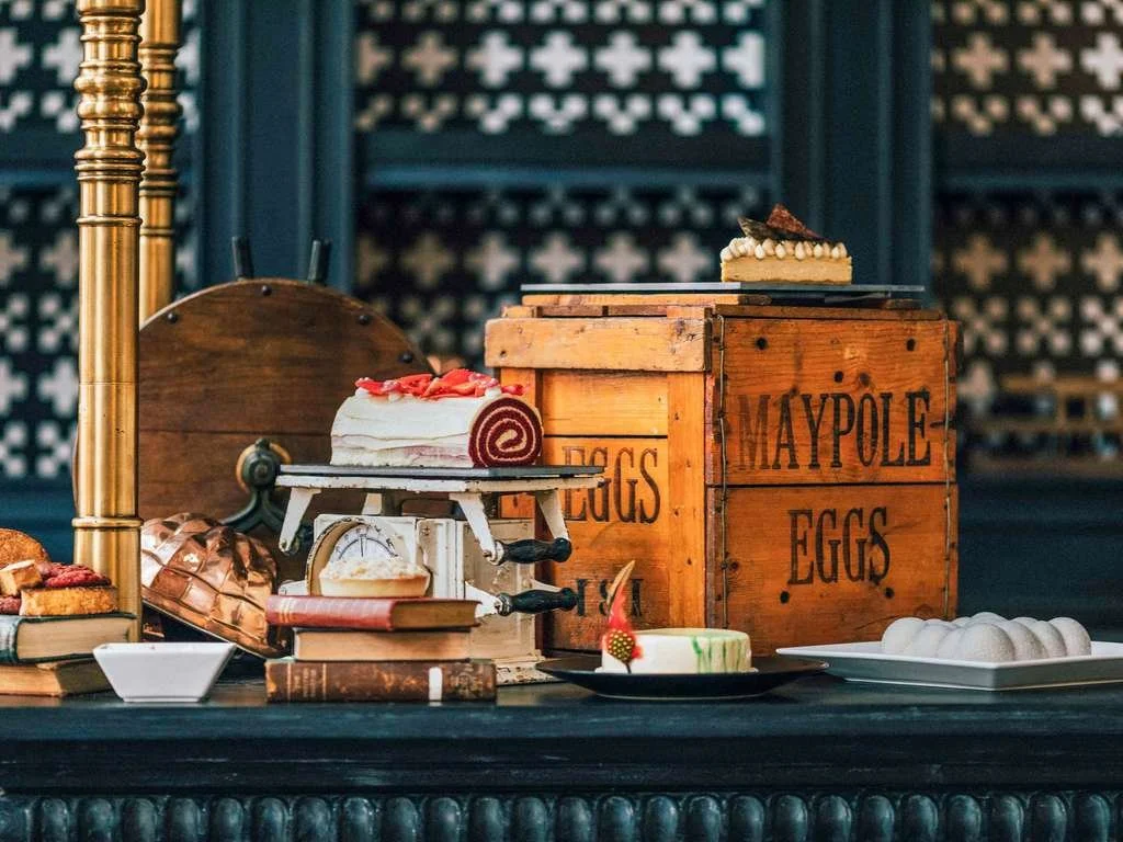 A display of desserts on a table, including a layered cake with strawberries on top, a pie or tart, and a plate with meringue cookies, set against a background with decorative panels and a wooden crate labeled "MAYPOLE EGG".