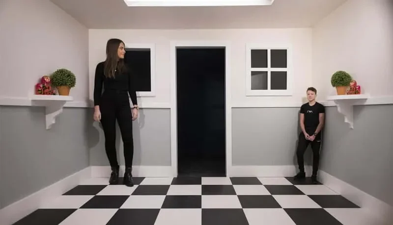 A woman and a man in a minimalist black and white room with checkered floor, three small windows, and potted plants on shelves, standing apart from each other.