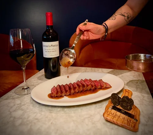 A person pouring sauce over sliced steak on a white oval plate, with a glass of red wine, a bottle of Château La Marzelle wine, and a wooden board with black truffles on a marble table.