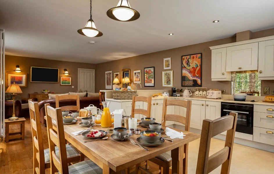 A kitchen and dining area with a wooden table set for breakfast, featuring bowls, mugs, juice, bread, and a pitcher, with various wall art and warm lighting.