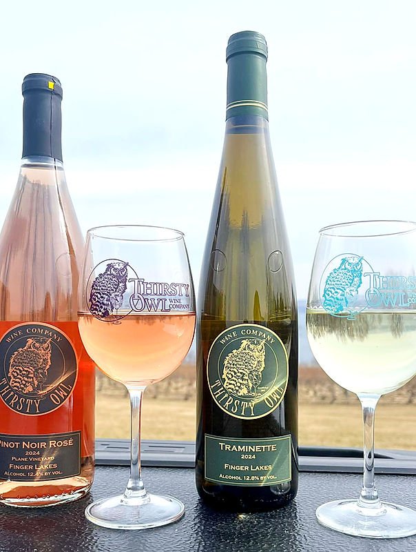 Two bottles of Thirsty Owl wine, one rosé and one white, with two wine glasses filled with rosé and white wine, outdoors with a field in the background.