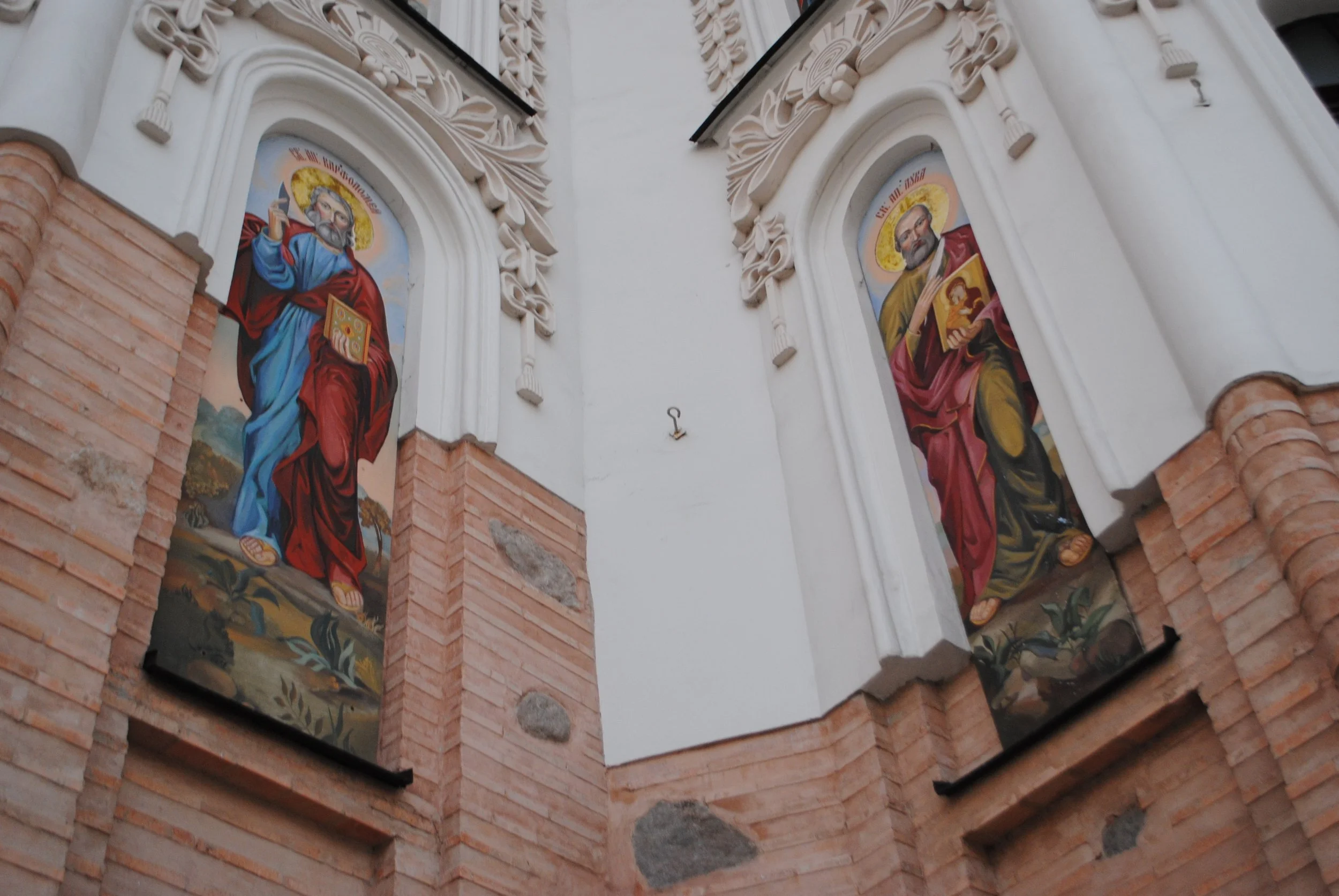 Paintings of saints or religious figures on the exterior of a church, framed by ornate white architectural details and brickwork.