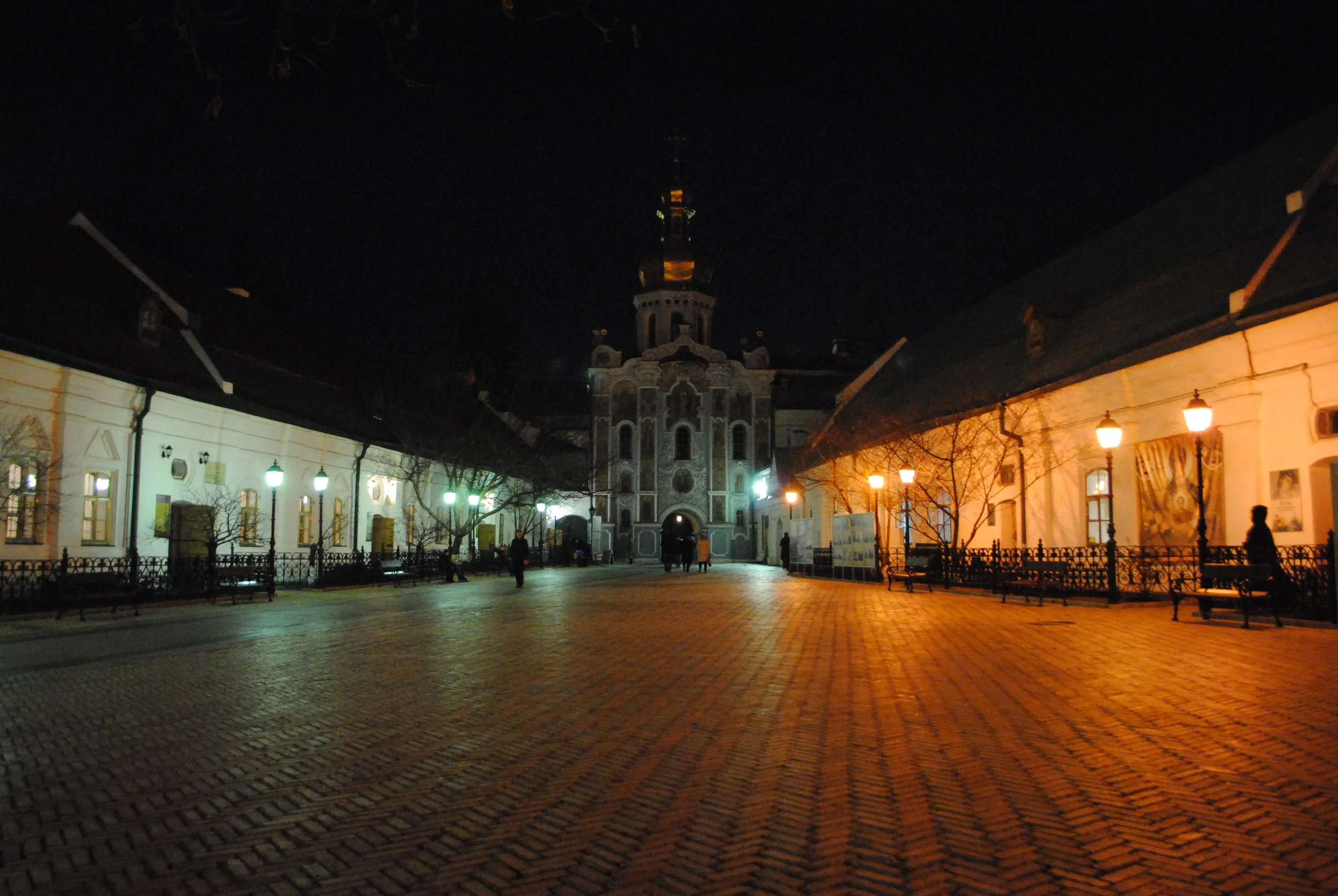 Night view of a historic church or cathedral with ornate architecture, illuminated by street lamps, surrounded by old buildings and benches in a square.