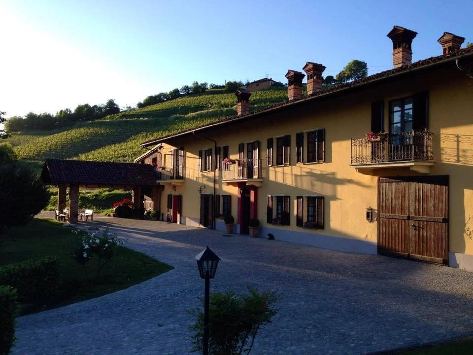 A large yellow house with black shutters and balconies, surrounded by greenery and a vineyard on a hillside, with a paved driveway and outdoor seating area in the foreground.