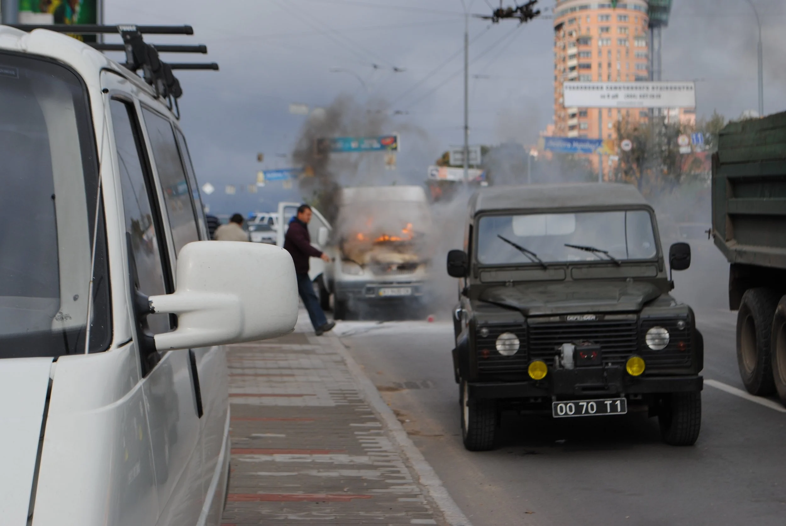 A street scene with multiple vehicles, including a white van in the foreground, a black vehicle, and a smoke-emitting vehicle in the background. There are buildings and street signs in the distance, with some smoke visible near a car that appears to 