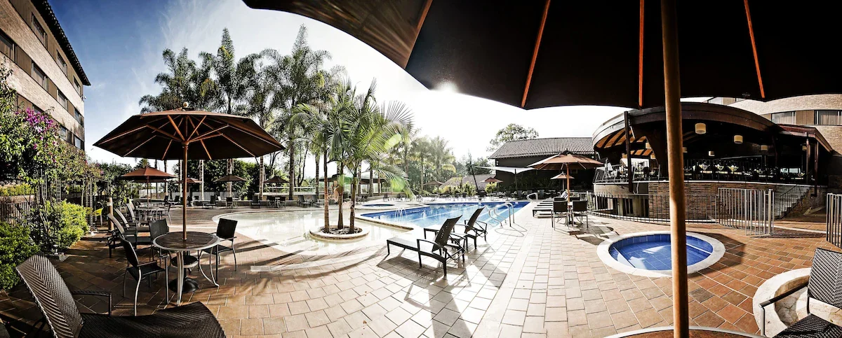 Pool area at a resort or hotel featuring lounge chairs, umbrellas, small hot tubs, palm trees, and a building in the background