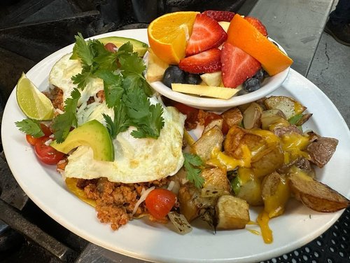 A plate of Mexican food with ground meat and toppings, fresh fruit salad, roasted potatoes, and garnishes including cilantro, avocado, lime, cherry tomatoes, and an egg on top of the seasoned meat