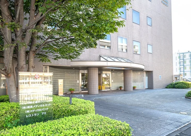 Exterior view of a modern multi-story hospital or medical building with a glass entrance, surrounded by green bushes and trees.