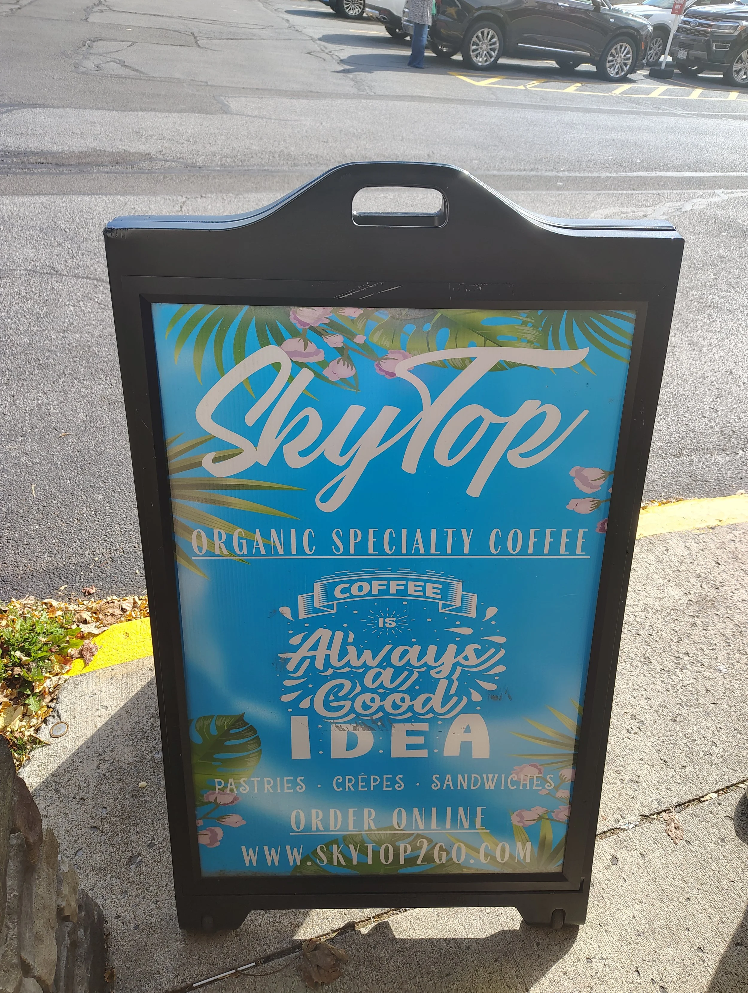 A black sidewalk sign advertising Sky Top Organic Specialty Coffee with a tropical design background, featuring text that says 'Coffee is Always a Good Idea' and mentions pastries, crepes, sandwiches, and online ordering at www.skytop2go.com.