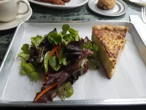 A slice of quiche accompanied by a side salad with mixed greens and shredded carrots on a metal tray