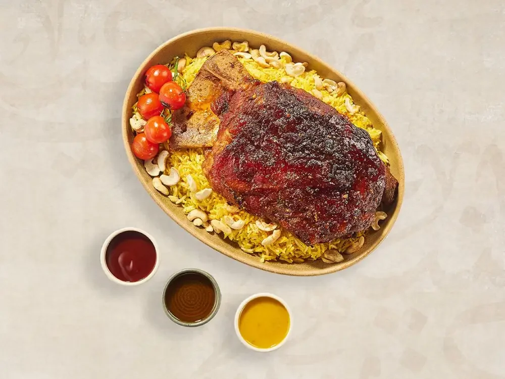 Bowl of cooked rice with marinated grilled beef, cherry tomatoes, cashews, and a side of three sauces.