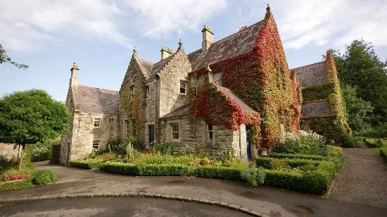 A large historic stone house covered with red and green ivy, surrounded by landscaped gardens and a curved driveway.