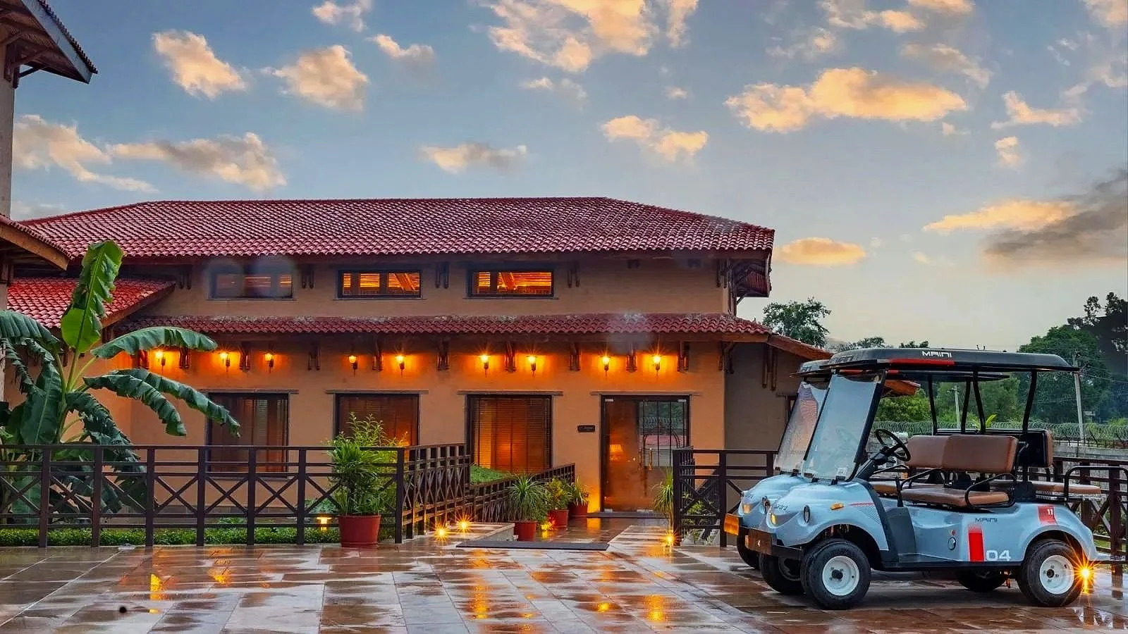 A two-story building with a red-tiled roof illuminated by warm lights, surrounded by potted plants, with a wet tiled patio in front. A golf cart is parked on the right side, and a blue sky with scattered clouds is seen overhead.