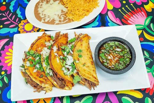 Three beef tacos with onions and cilantro on a rectangular white plate, with a side of salsa in a small black bowl. In the background, there is a plate with refried beans topped with melted cheese and Spanish rice. The tablecloth has vibrant floral patterns.