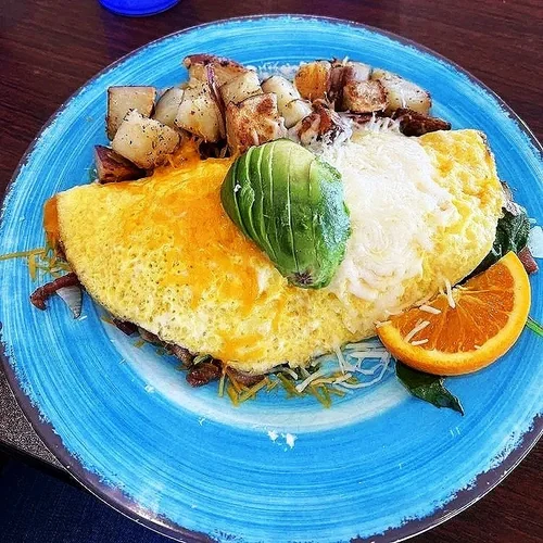 Omelette topped with shredded cheese, sliced avocado, and a dollop of sour cream, served with roasted potatoes, lettuce, and a slice of orange on a blue plate.