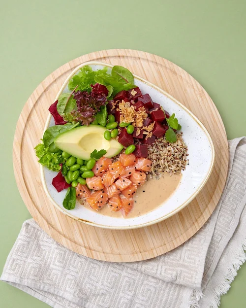 A white bowl with mixed salads, diced raw fish, rice, avocado, and dressing on a round wooden plate with a white cloth underneath on a green background.