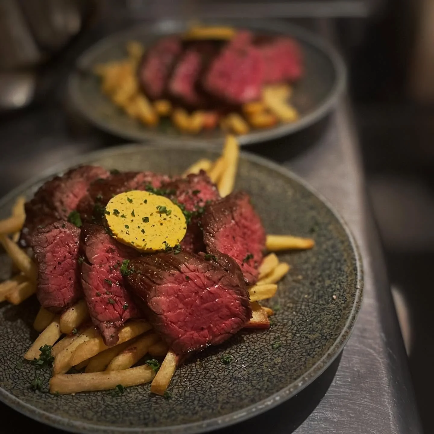 A plate of cooked steak slices on top of French fries, topped with a pat of herb butter and garnished with chopped herbs. In the background, another plate with more steak slices and fries is visible.