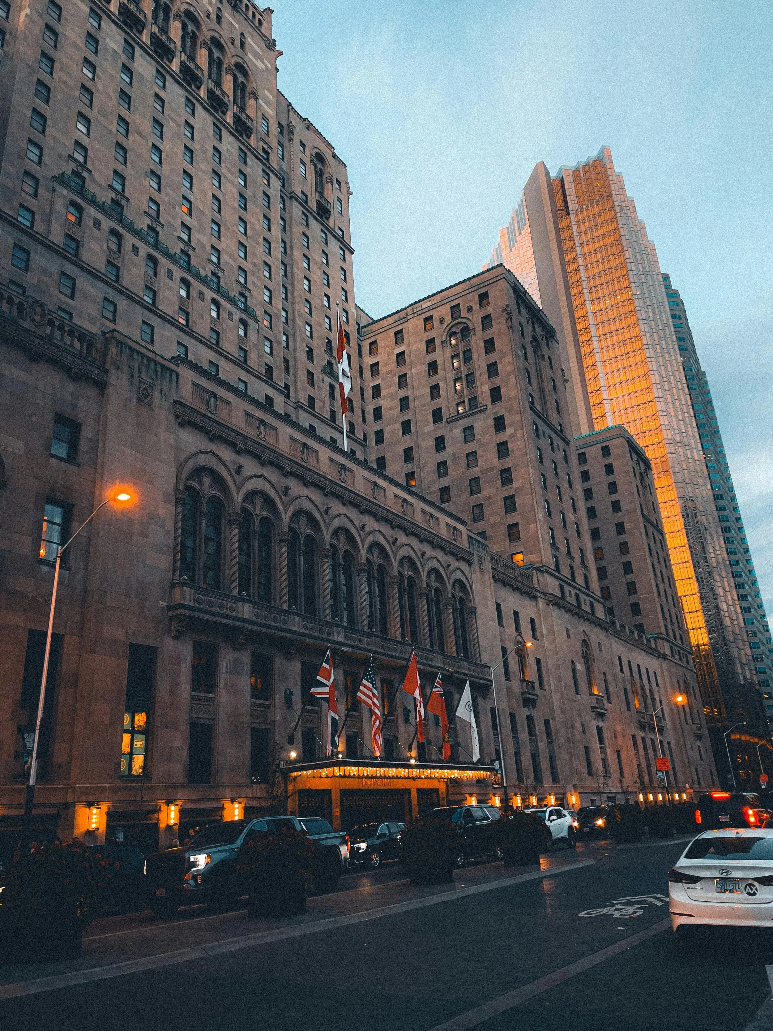 City street with tall buildings, parked cars, and flags outside a building with illuminated signage, during dusk.